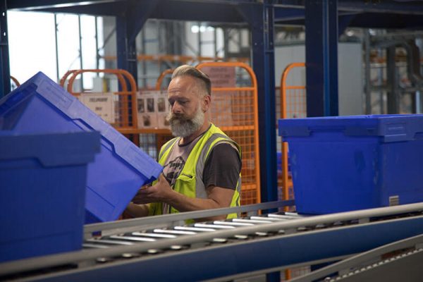 Horner casual warehouse pick packer worker in hi-vis vest lifting blue box off conveyor belt